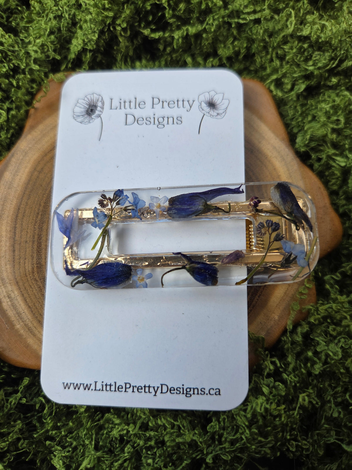 A clear acrylic hair claw clip with embedded blue flowers and green leaves, displayed on a white branded card over a wood slice and moss background.