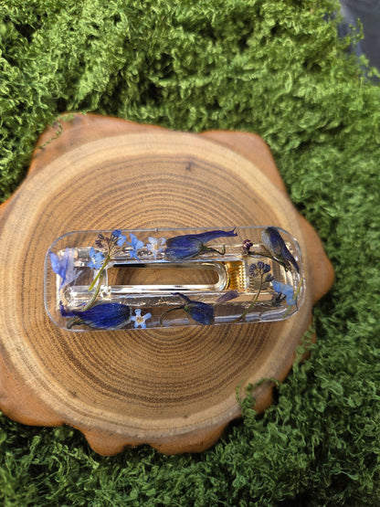 A clear acrylic hair claw clip containing blue and white wildflowers, displayed on a circular wood slice surrounded by green moss.